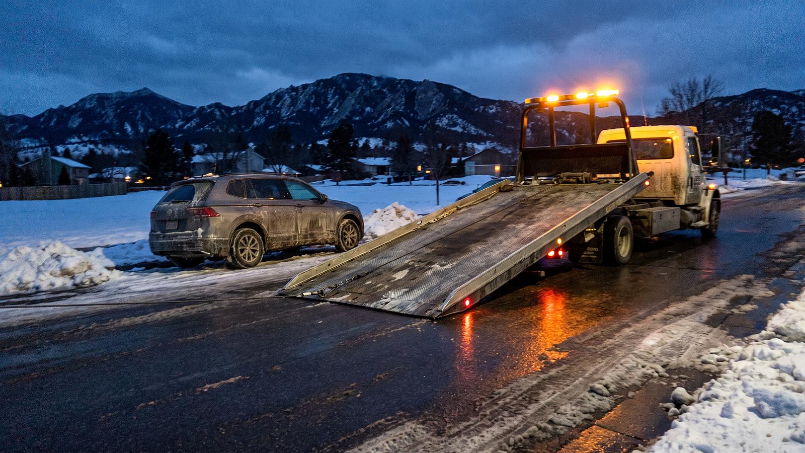A flatbed tow truck with its bed tilted to the road at dusk, an SUV waiting at the curb and the Flatirons behind